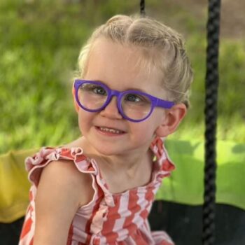 Drug repurposing is a promising area in research on mitochondrial disease. In this image, a blond girl, named Millie, with big purple glasses smiles at the camera while sitting on a trampoline on a sunny day.