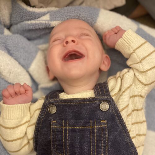 Smiling baby boy named Brooks, affected by Leigh syndrome, lays on a blanket with overalls on. This image links to Brooks’s story about his life and diagnosis.