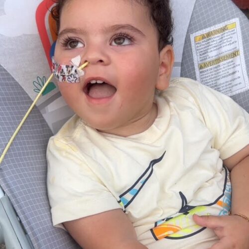 Young boy named Aiden, affected by Leigh syndrome, sits in a chair wearing a cream-colored shirt with a shark design. He has a medical tube secured with a decorative sticker on his face and looks upward with a joyful expression. This image links to Aiden’s story about his life and diagnosis.