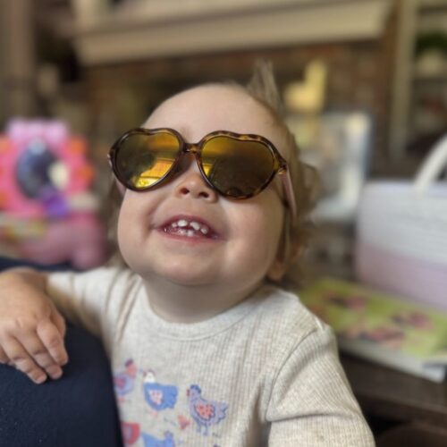 Young child named AJ, affected by Leigh syndrome, smiles brightly while wearing heart-shaped sunglasses. AJ is dressed in a light shirt with colorful bird designs, leaning with joy in a cozy indoor setting. This image links to AJ’s story about life and diagnosis.