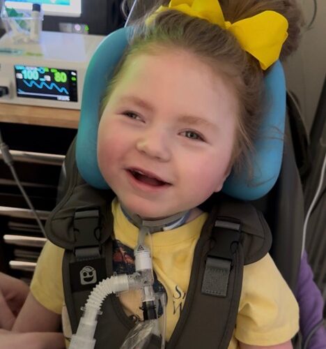 Smiling young girl named Brighton with a bright yellow bow in her hair, seated in a supportive medical chair with breathing equipment. She is living with Leigh syndrome, and this image links to her personal story about her life and diagnosis.