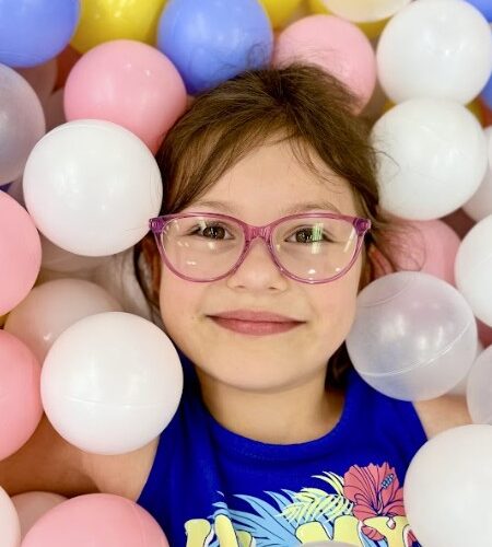 Young girl named Julia, affected by ATP6 mitochondrial disease Leigh syndrome, smiles while surrounded by colorful plastic balls in shades of white, pink, blue, and yellow. She wears glasses and a bright blue shirt with a floral design. This image links to Julia’s story about her life and diagnosis.