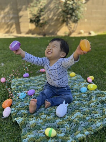 Young boy named Kenji, affected by Leigh syndrome, sits on a crocheted blanket outdoors surrounded by colorful Easter eggs. He is smiling with his eyes closed and arms raised joyfully, holding two eggs in his hands. This image links to Kenji’s story about his life and diagnosis.