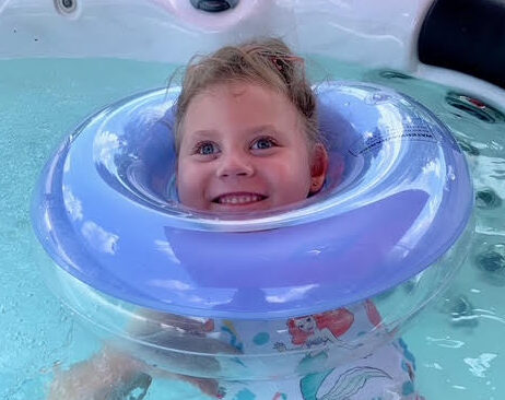 Young girl named Lucinda, who was affected by Leigh syndrome and has since passed away, smiles brightly while floating in the water with a supportive purple swim ring. She wears a swimsuit with mermaid designs. This image links to Lucinda’s story about her life, diagnosis, and legacy.