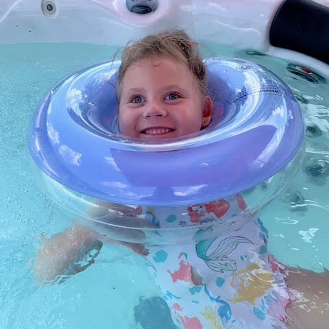 Young girl named Lucinda, who was affected by Leigh syndrome and has since passed away, smiles brightly while floating in the water with a supportive purple swim ring. She wears a swimsuit with mermaid designs. This image links to Lucinda’s story about her life, diagnosis, and legacy.